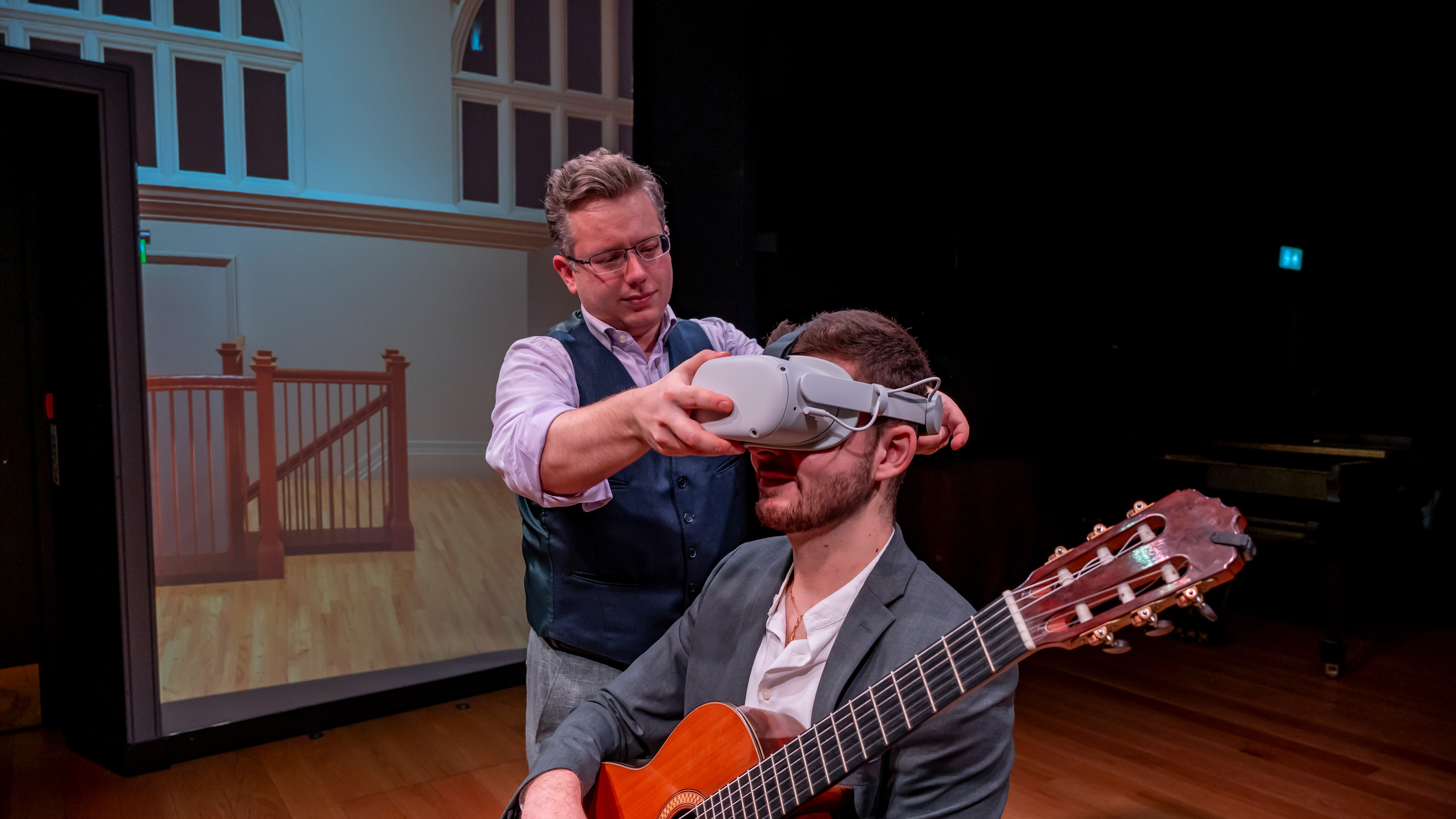 A man wearing a waist coat and suit putting on a VR headset on a male student in a suit, holding a guitar.